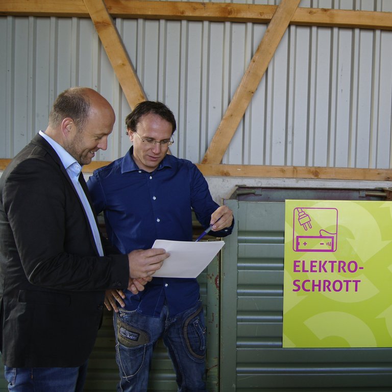 zwei Männer beraten sich bezüglich einer Liste vor einem Elektroschrott-Container